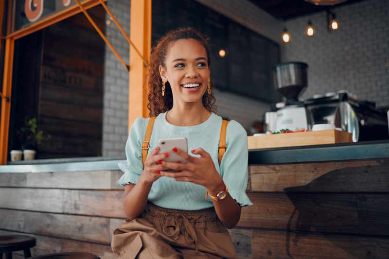 Eine junge Frau mit Locken sitzt auf einer Bank vor einem Café und überlegt sich eine Google Bewertung, die sie auf ihr Smartphone eingeben möchte